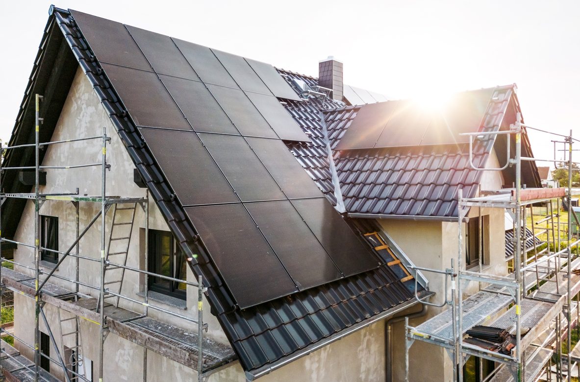 The roof of a German single-family home under construction. Photographed at sunrise with a lens flare from the rising sun. Prominent dark solar panels are mounted on the tiled gable roof.